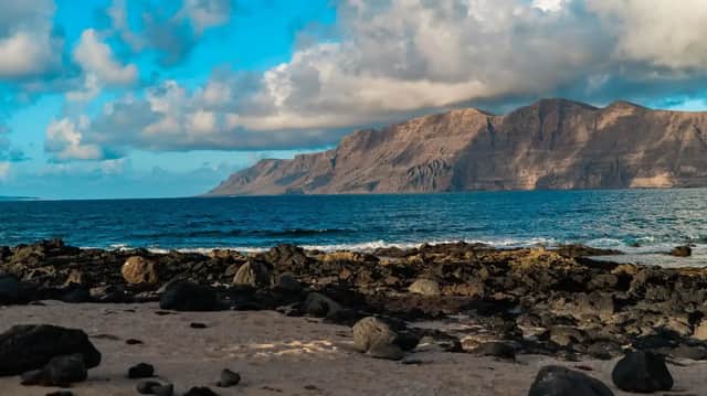 Una costa rocciosa, con sabbia e pietre vulcaniche, si affaccia su una grande catena montuosa oltre l'oceano blu, sotto un cielo parzialmente nuvoloso.