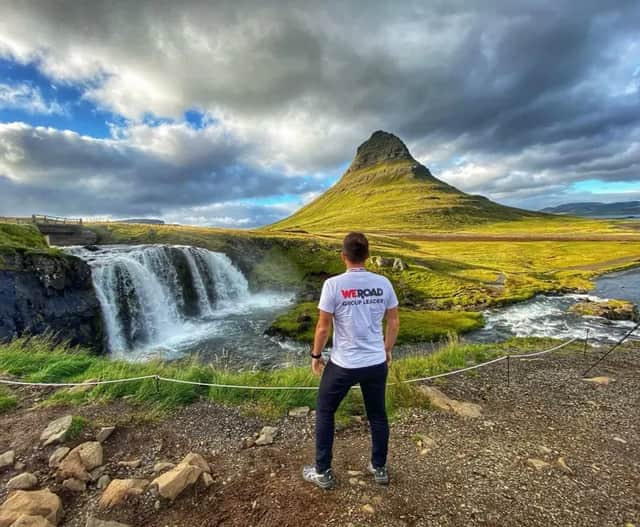 Un líder de grupo WeRoad visto de espaldas, mirando una cascada que fluye junto a una gran montaña verde bajo un cielo nublado.