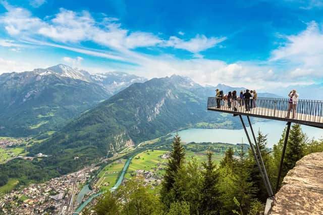 A WeRoad group trip stands on a suspended viewing platform, admiring a panoramic view of a mountain valley, a lake, and a town.