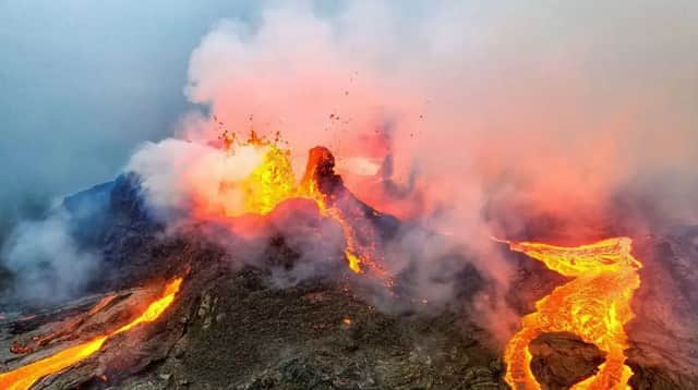 Un volcan actif entre en éruption, avec de la lave en fusion s'écoulant en rivières le long de son flanc sombre et rocheux sous un ciel rempli de fumée.