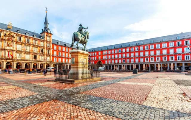 A bronze equestrian statue stands in the center of a large cobblestone plaza, surrounded by historic red buildings as people walk by.