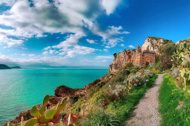 Ein Feldweg führt zu einem Steinmonument auf einer grünen Klippe mit Blick auf ein türkisfarbenes Meer unter einem bewölkten Himmel.