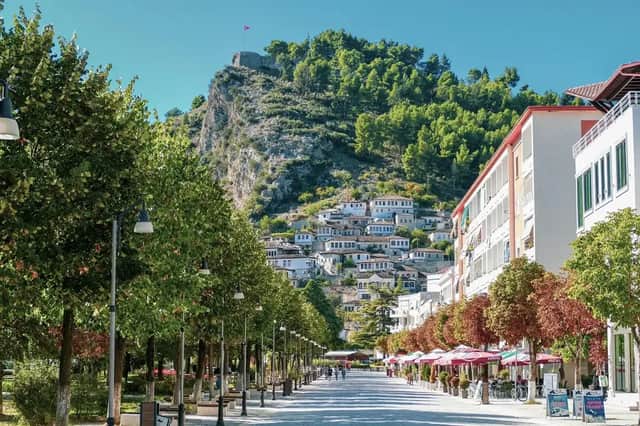 Una calle ancha y arbolada conduce a un pueblo en la ladera con casas blancas y una fortaleza en la cima.