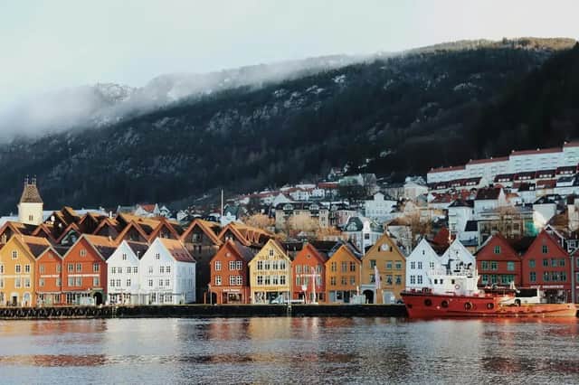 Colorful wooden houses line a waterfront with a red boat moored nearby, at the base of a forested, misty hill.