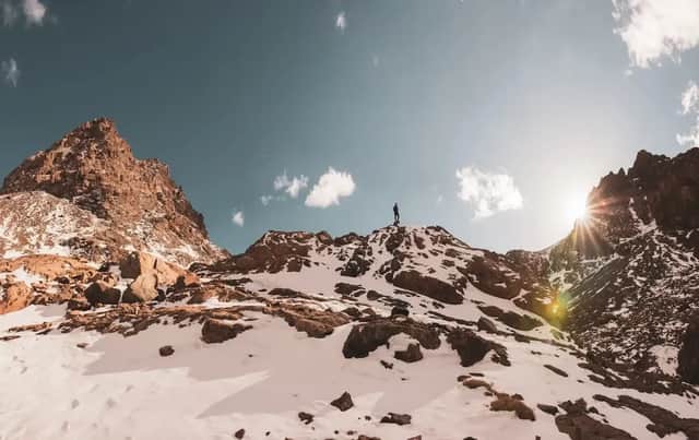 Une silhouette se découpe sur le ciel, sur un sommet montagneux enneigé et rocheux, sous un soleil éclatant.