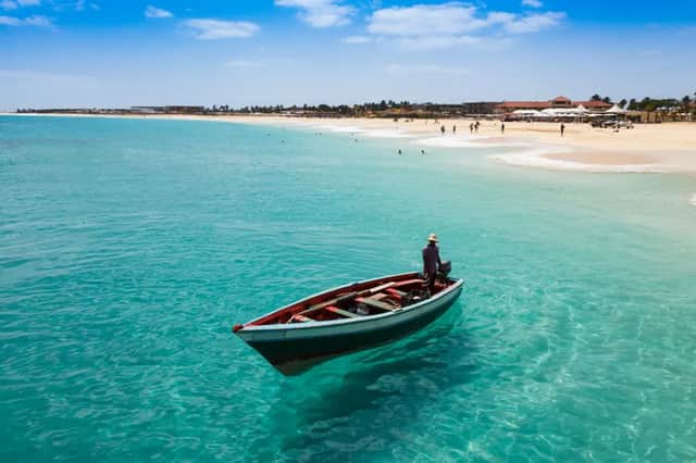 Une personne coiffée d'un chapeau se tient dans un petit bateau sur une eau turquoise et claire, avec une plage de sable et des gens à l'arrière-plan.