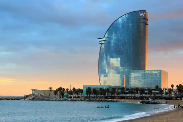 A modern, sail-shaped glass hotel stands by a sandy beach at sunset, with palm trees and people paddleboarding in the sea.