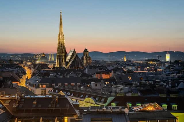 Una guglia gotica illuminata svetta su un paesaggio urbano al tramonto, con montagne lontane sotto un cielo crepuscolare dai colori vivaci.
