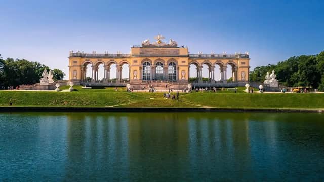 Un gran edificio ornamentado con arcos se alza sobre una colina verde, reflejado en un gran estanque en un día claro y soleado.