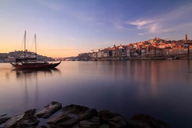 Una barca de madera tradicional en un río tranquilo al atardecer, con los coloridos edificios de una ciudad en la ladera en la orilla opuesta, bajo un cielo rosado y azul.