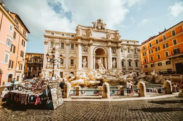 An ornate white marble fountain in a city square on a sunny day, with a souvenir stall on the cobblestone foreground.