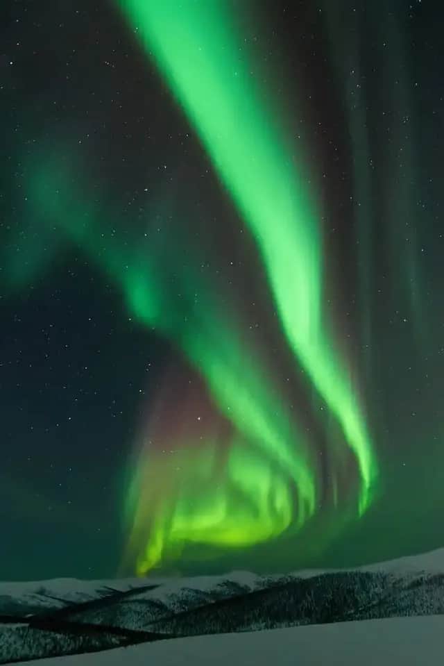 Helle grüne Bänder des Nordlichts leuchten am sternenklaren Nachthimmel über einer verschneiten Bergkette.