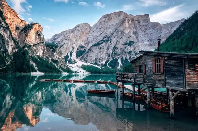 A wooden boathouse on stilts and rowboats on a calm lake, with snow-capped mountains reflected in the water.