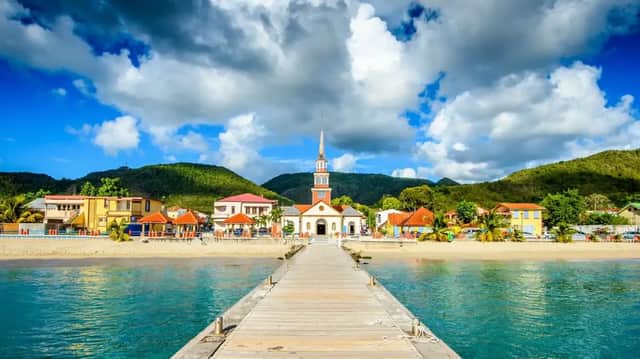 Une jetée en bois s'avance sur des eaux turquoise vers une ville côtière avec une église, adossée à des collines verdoyantes sous un ciel nuageux.