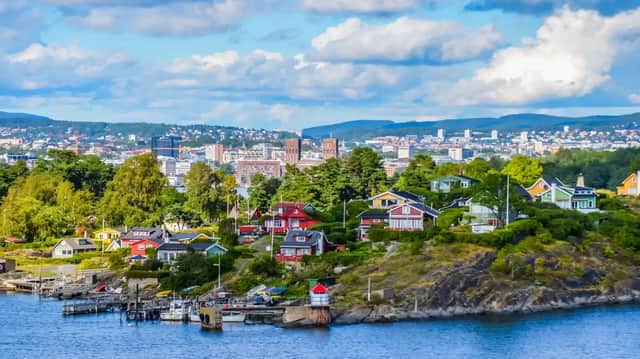 Des maisons colorées parsèment un littoral vert et rocheux devant une ligne d'horizon urbaine lointaine sous un ciel partiellement nuageux.