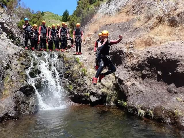 Una persona de un viaje en grupo de WeRoad, con traje de neopreno y casco, se lanza a una piscina natural junto a una cascada mientras otros observan.
