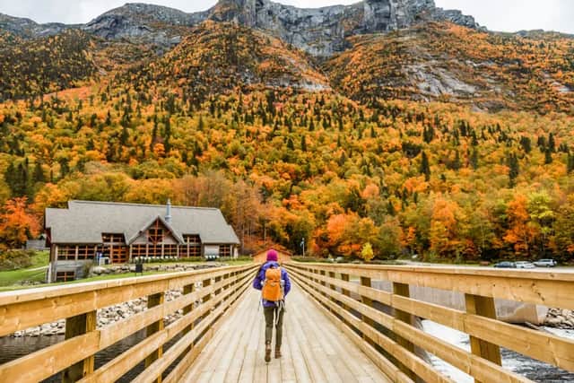A person with a backpack walks across a wooden bridge towards a large lodge and a mountain covered in colorful autumn foliage.