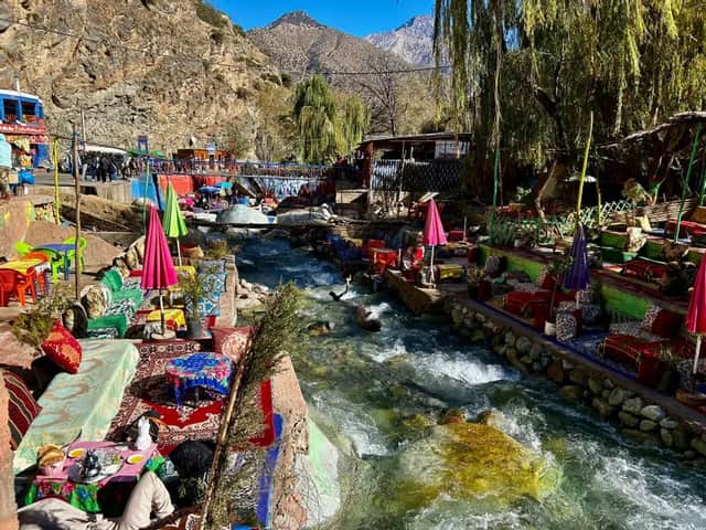 Coloridas cafeterías al aire libre con vibrantes zonas de asientos y sombrillas bordean las orillas de un río caudaloso al pie de una montaña soleada.
