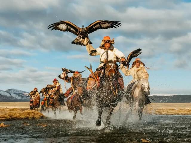 Cacciatori di aquile, in abiti tradizionali, galoppano a cavallo attraverso un fiume, con le aquile al braccio.