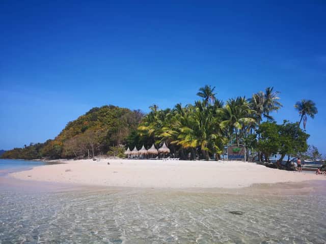 Vista dall'acqua limpida e poco profonda di un'isola tropicale con spiaggia di sabbia bianca, palme e una collina boscosa sotto un cielo blu.