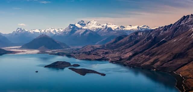 Vue aérienne d'un grand lac bleu avec de petites îles, entouré de montagnes escarpées et de sommets enneigés en arrière-plan.