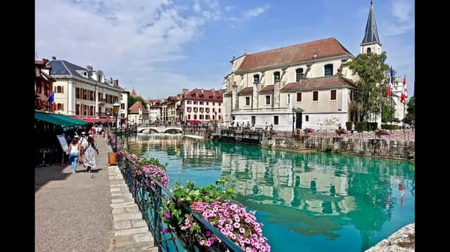 Un canal turquoise traverse une ville historique, avec des gens se promenant sur la promenade au bord de l'eau et des bâtiments se reflétant dans l'eau.