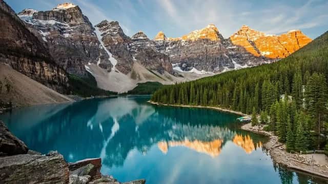 Picos de montañas nevados y soleados que se reflejan en un lago turquesa en calma, rodeado por un denso bosque de pinos.