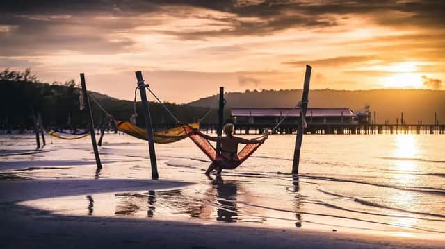 Une personne vue de dos est assise dans un hamac rouge au-dessus de l'eau sur une plage, lors d'un coucher de soleil doré.