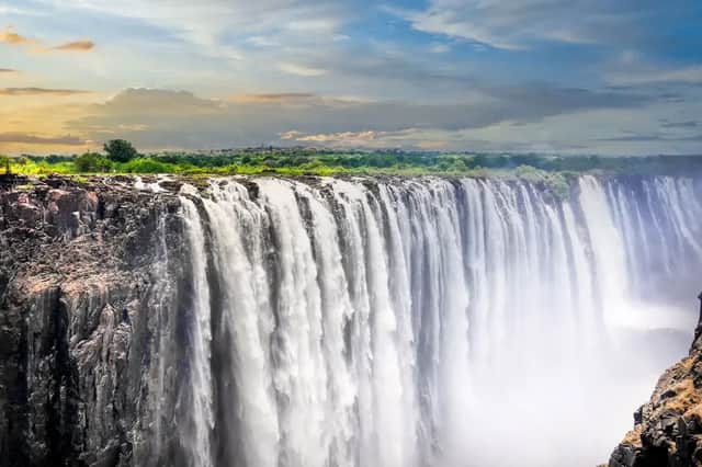 A massive waterfall cascades over a wide, rocky cliff face, with a line of green trees in the background under a partly cloudy sky.