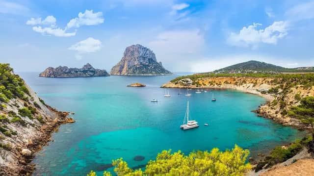 Veleros anclados en una cala turquesa rodeada de acantilados rocosos, con una gran isla visible a lo lejos bajo un cielo parcialmente nublado.