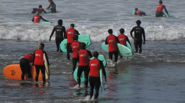 A WeRoad group trip wearing wetsuits carries surfboards into the ocean for a lesson with their coaches.
