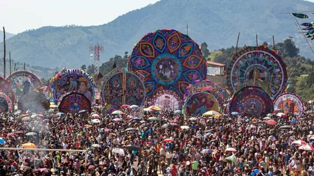 Eine riesige Menschenmenge versammelt sich auf einem Open-Air-Festival, um mehrere riesige, bunte, verzierte Drachen zu bestaunen, die vor einer Bergkette ausgestellt sind.