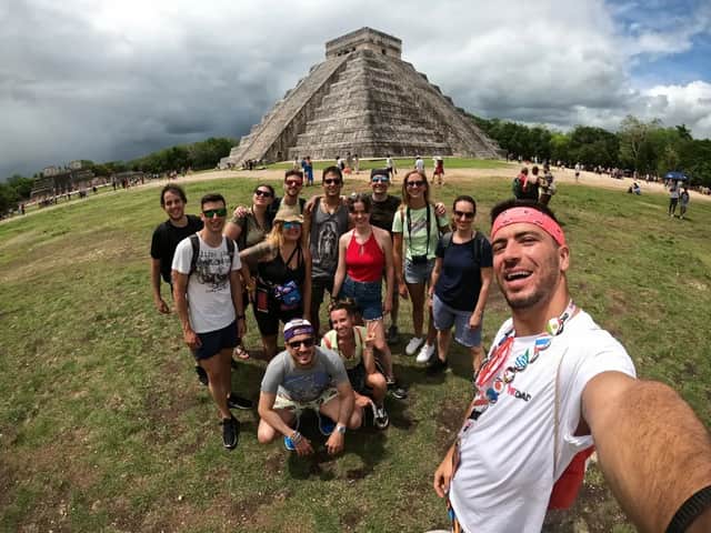 Un viaje en grupo de WeRoad se toma una selfie sonriente en un campo de hierba frente a una gran pirámide de piedra antigua bajo un cielo nublado.