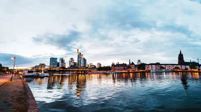 Una vista grandangolare dello skyline di una città al tramonto, con edifici illuminati che si riflettono nel fiume sottostante sotto un cielo nuvoloso.