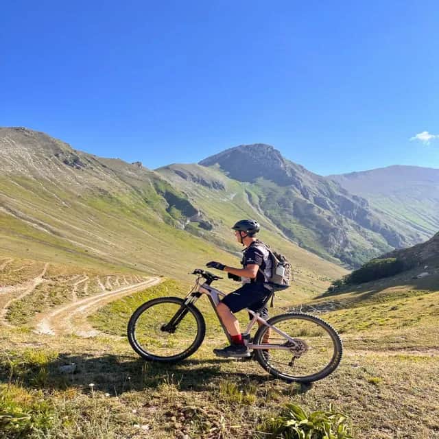 Un mountain biker con il casco si ferma su un pendio erboso, ammirando il panorama di una catena montuosa verde.