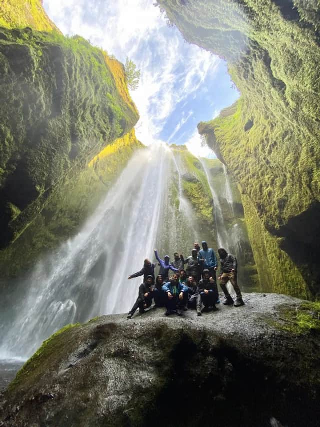 A WeRoad group trip poses on a large rock at the base of a waterfall inside a mossy canyon.