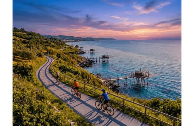 Veduta aerea di un viaggio di gruppo WeRoad in bicicletta su un sentiero costiero tortuoso con tradizionali moli da pesca sul mare al tramonto.