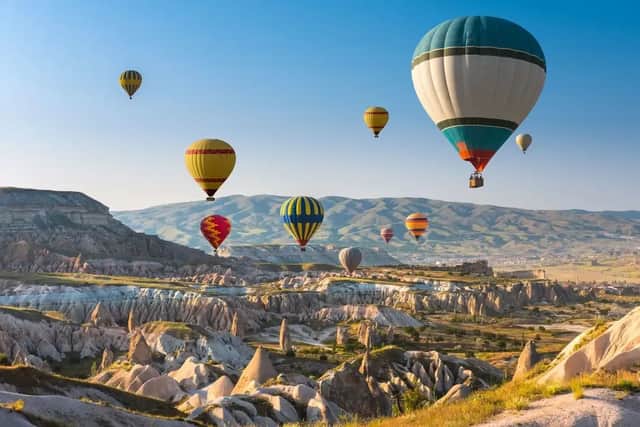 Molte mongolfiere colorate che sorvolano un vasto paesaggio roccioso con formazioni geologiche uniche, sotto un cielo azzurro e limpido.