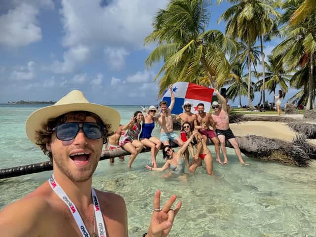 Un homme prend un selfie avec son groupe WeRoad, posant sur un palmier couché dans une eau tropicale cristalline, tenant un drapeau panaméen.