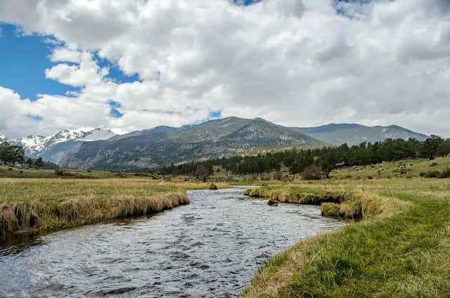 A river flows through a grassy meadow towards a range of snow-capped mountains under a partly cloudy sky.