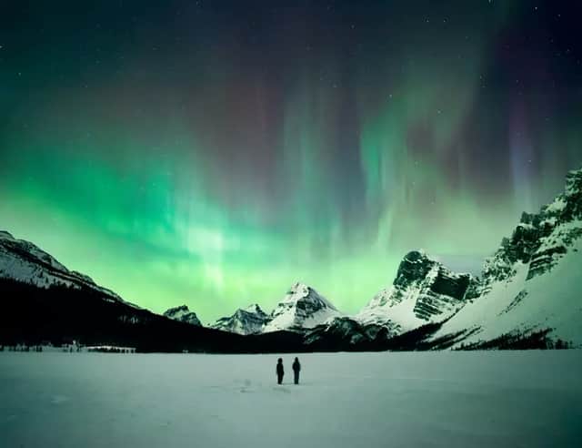 Two people stand on a vast snowy landscape watching the green and purple aurora borealis over snow-capped mountains.