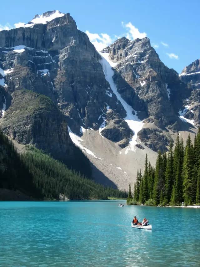 A WeRoad group trip paddles a canoe on a turquoise alpine lake surrounded by pine forests and snow-capped mountains.
