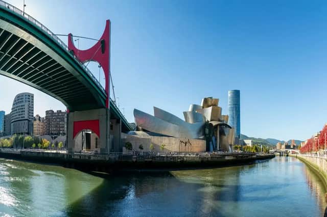 A panoramic view of a modern metallic museum and a red bridge over a river on a sunny day.