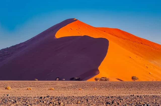 A large orange sand dune casts a deep shadow across itself in a rocky desert landscape under a clear blue sky.
