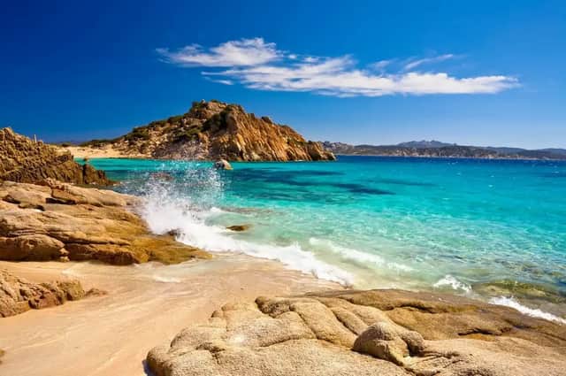 A wave splashes against the rocks of a sandy cove with turquoise water under a bright blue sky.