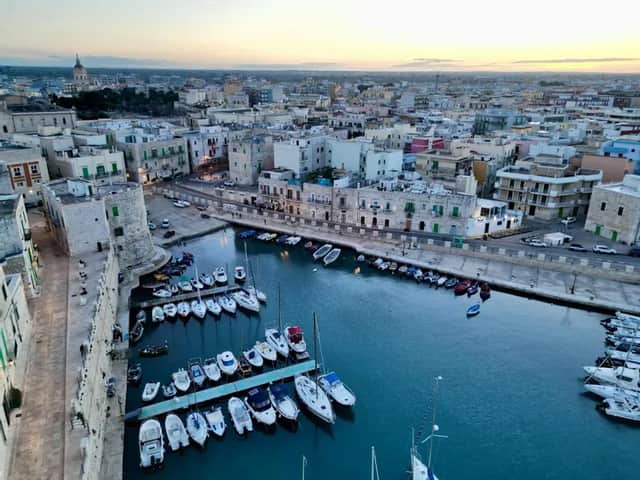 Una vista aerea di una città costiera al tramonto, con molte barche ormeggiate in un piccolo porto accanto a una storica torre di pietra.