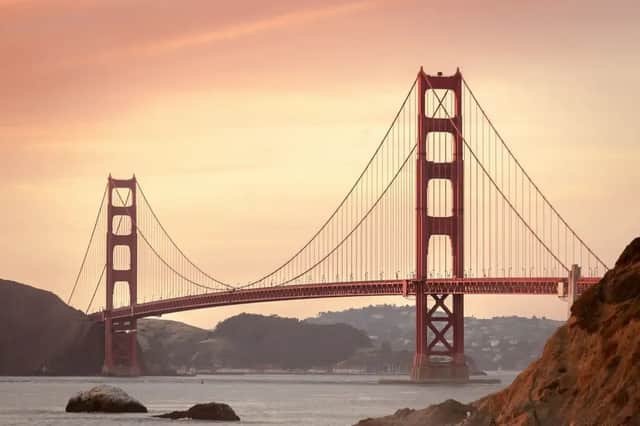 Eine große rote Hängebrücke überspannt ein Gewässer mit Hügeln im Hintergrund unter einem warm getönten Himmel bei Sonnenuntergang.