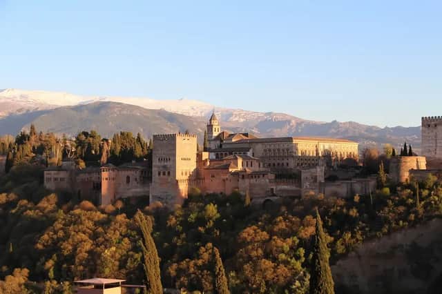 Una grande fortezza in pietra su un fianco di collina boscoso, illuminata da una luce dorata, con montagne innevate sullo sfondo.