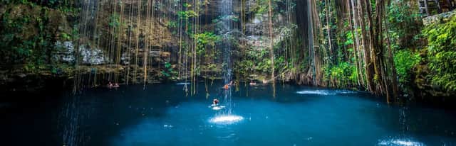 Un groupe WeRoad en voyage nage dans l'eau bleue profonde d'un cénote, avec une cascade et de longues lianes suspendues aux falaises.