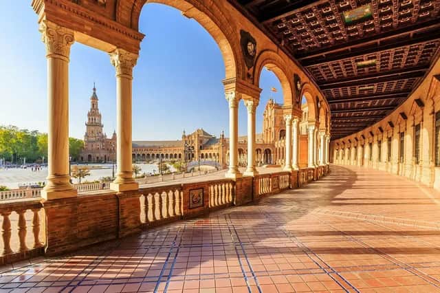 Vue d'une arcade ensoleillée avec colonnes et arches, surplombant une grande place et un bâtiment historique.